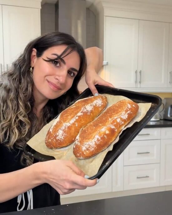 Woman holding a baking sheet with two golden-brown French baguettes; a delicious French baguette recipe.