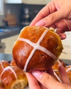 Close-up of a golden-brown hot cross bun held by hands, featuring a white cross. Quick hot cross bun recipe.