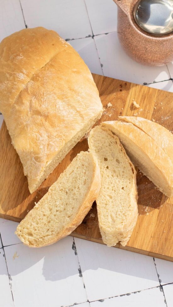 Sliced Italian sandwich bread loaf on a wooden cutting board with a pink teapot.