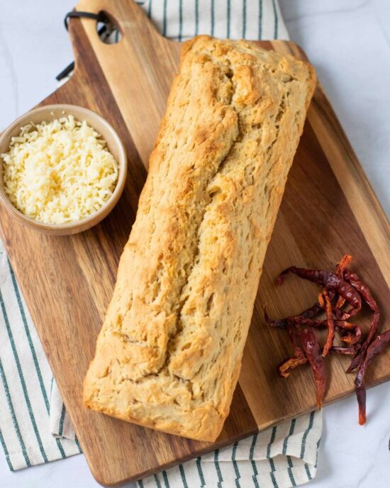 Golden beer bread loaf on a wooden board with shredded cheese and dried chili peppers. Simple beer bread recipe.