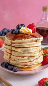 Stack of diner style pancakes with butter, strawberries, blueberries, and syrup.