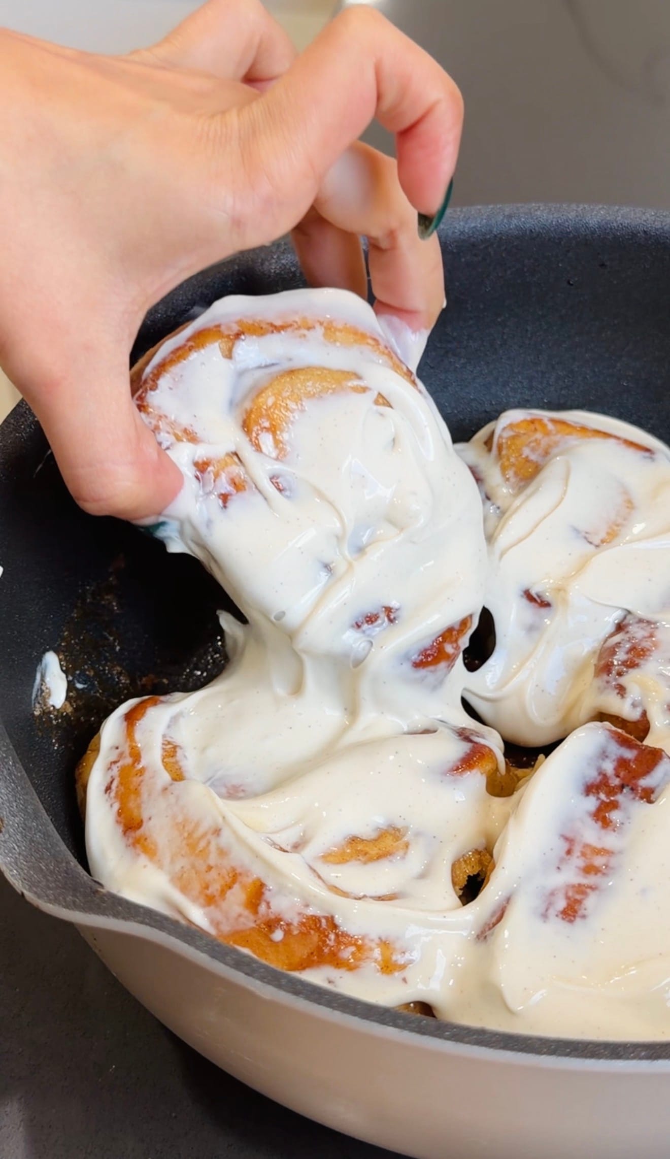 Hand lifting a frosted cinnamon roll from a pan of freshly baked rolls.
